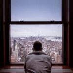 View of the Manhattan Skyline from behind a window. A man is looking out of the window with his back to the camera.